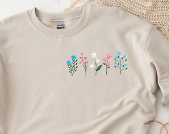 May include: A beige sweatshirt with a floral design featuring pink, blue, and white flowers. The flowers are arranged in a row across the chest of the sweatshirt.