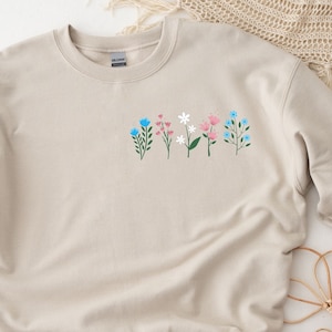 May include: A beige sweatshirt with a floral design featuring pink, blue, and white flowers. The flowers are arranged in a row across the chest of the sweatshirt.