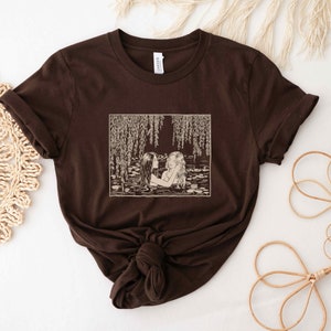 May include: A brown t-shirt with a vintage illustration of two women swimming in a pond with willow trees hanging over the water. The illustration is in a cream color.