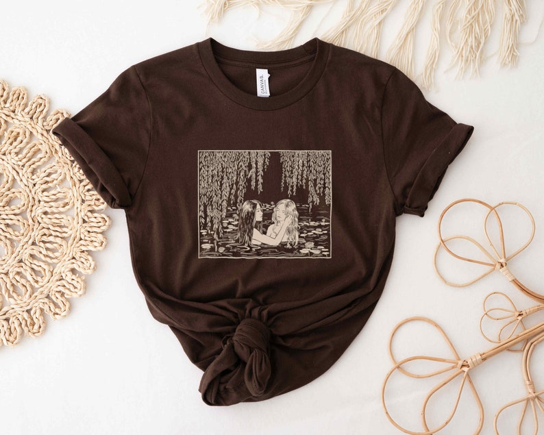 May include: A brown t-shirt with a vintage illustration of two women swimming in a pond with willow trees hanging over the water. The illustration is in a cream color.