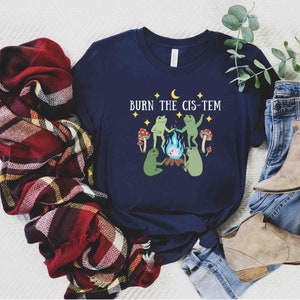 Puede incluir: Una camiseta azul marino con un gr&aacute;fico de dos ranas alrededor de una fogata con el texto "BURN THE CIS-TEM" encima.