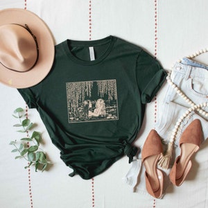 May include: A dark green t-shirt with a vintage illustration of two women in a pond with willow trees. The illustration is in a cream color.