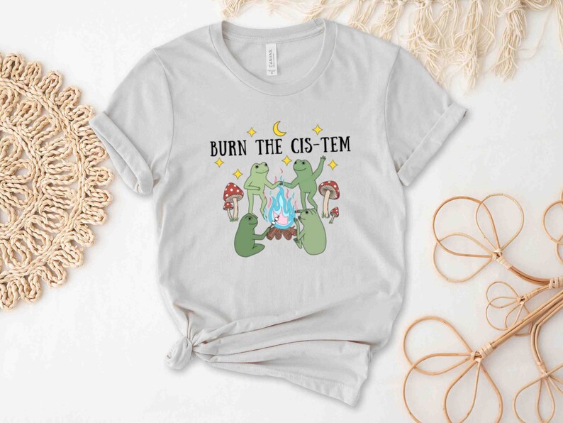 Puede incluir: Una camiseta gris claro con el texto "BURN THE CIS-TEM" impreso en ella. El dise&ntilde;o presenta cuatro ranas alrededor de una fogata con setas y estrellas.