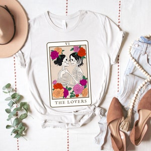 Peut inclure: T-shirt blanc avec un motif de la carte de tarot des Amoureux repr&eacute;sentant deux squelettes qui s'embrassent. La carte est entour&eacute;e de fleurs color&eacute;es et le texte "VI LES AMOUREUX" est imprim&eacute; sous l'image.