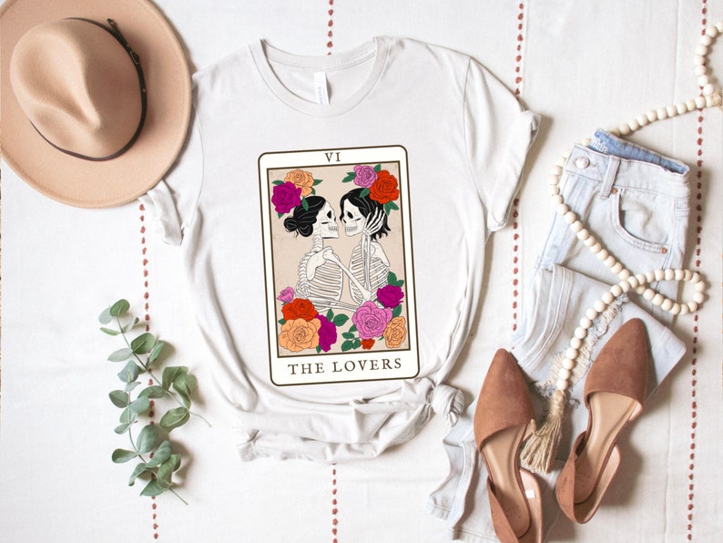 Peut inclure: T-shirt blanc avec un motif de la carte de tarot des Amoureux repr&eacute;sentant deux squelettes qui s'embrassent. La carte est entour&eacute;e de fleurs color&eacute;es et le texte "VI LES AMOUREUX" est imprim&eacute; sous l'image.