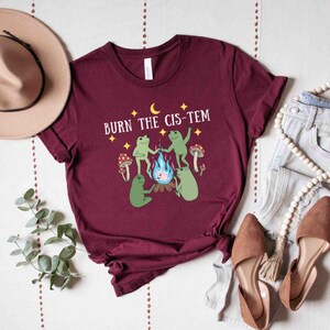 Puede incluir: Una camiseta color burdeos con un gr&aacute;fico de tres ranas alrededor de una fogata con el texto "BURN THE CIS-TEM" encima.