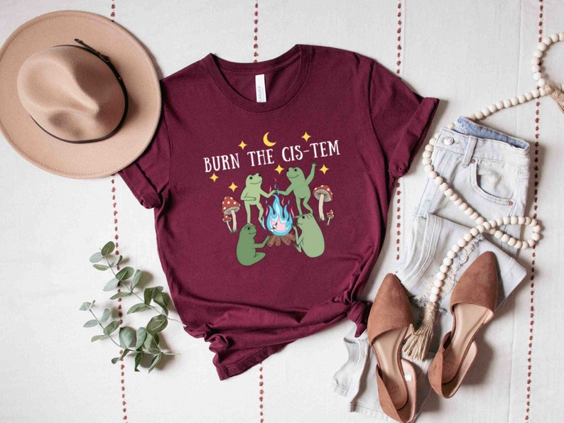 Puede incluir: Una camiseta color burdeos con un gr&aacute;fico de tres ranas alrededor de una fogata con el texto "BURN THE CIS-TEM" encima.