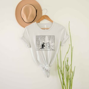 May include: A light gray t-shirt with a black and white illustration of two people standing in a forest. The illustration is surrounded by a white border.