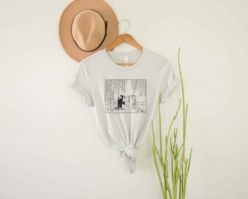 May include: A light gray t-shirt with a black and white illustration of two people standing in a forest. The illustration is surrounded by a white border.