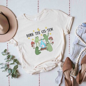 Puede incluir: Una camiseta blanca con un gr&aacute;fico de tres ranas alrededor de una fogata con el texto "BURN THE CIS-TEM" encima.