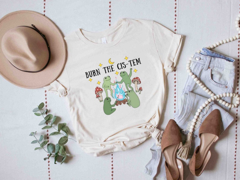 Puede incluir: Una camiseta blanca con un gr&aacute;fico de tres ranas alrededor de una fogata con el texto "BURN THE CIS-TEM" encima.