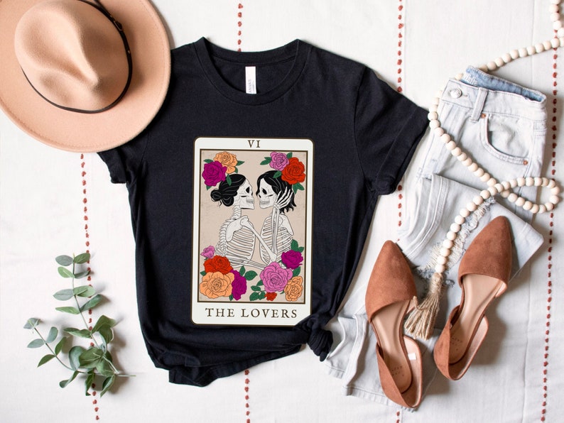 Peut inclure: T-shirt noir avec un motif de carte de tarot repr&eacute;sentant deux squelettes qui s'embrassent. La carte est intitul&eacute;e "The Lovers" et porte le chiffre romain VI en haut.