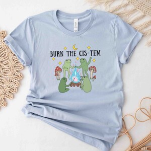 Puede incluir: Camiseta azul claro con un gr&aacute;fico de tres ranas alrededor de una fogata con el texto "BURN THE CIS-TEM" en negro.