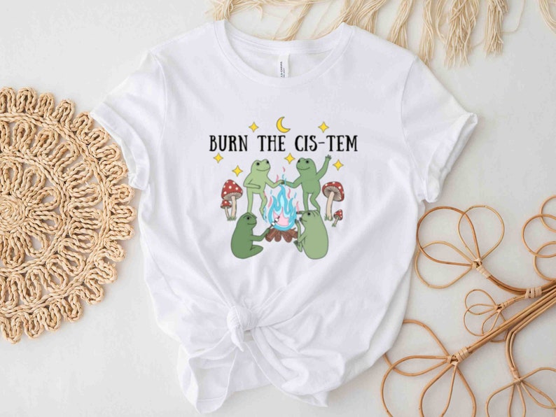 Puede incluir: Camiseta blanca con un gr&aacute;fico de cuatro ranas alrededor de una fogata con el texto "BURN THE CIS-TEM" encima.