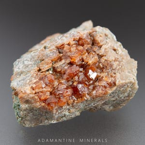 May include: A close-up of a rough, brown and gray rock with a cluster of small, orange-red crystals. The crystals are clustered together in the center of the rock.