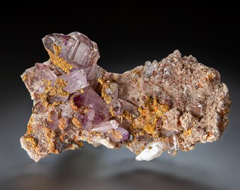May include: A cluster of translucent, light purple amethyst crystals embedded in a rough, textured matrix of brown and tan rock. The mineral specimen features sharp, pointed crystal formations with visible internal facets and earthy deposits.
