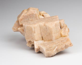 May include: A cluster of pale beige, blocky mineral crystals with a matte, textured surface. The specimen features multiple interlocking cubic and rectangular formations with visible striations and natural edges, creating a dense, geometric structure.