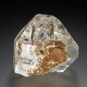 May include: A clear, multifaceted quartz crystal specimen featuring a prominent, textured patch of reddish-brown iron oxide inclusions embedded within its transparent, geometric structure, showcasing natural internal fractures and sharp edges.