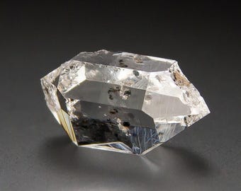 May include: A clear, double-terminated quartz crystal featuring natural facets and internal inclusions of dark, opaque mineral specks. The stone exhibits a high degree of transparency with sharp, geometric edges and a polished, reflective surface.