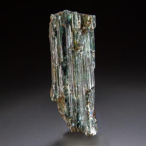 May include: A raw, elongated tourmaline crystal featuring vertical striations and a translucent, multi-faceted structure. The mineral displays a gradient of deep forest green, teal, and subtle golden-brown hues with a reflective, glassy surface.