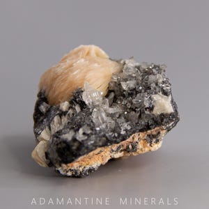 May include: A cluster of light pink barite crystals on a dark gray matrix. The barite crystals are translucent and have a pearly luster. The matrix is covered in small, clear quartz crystals.
