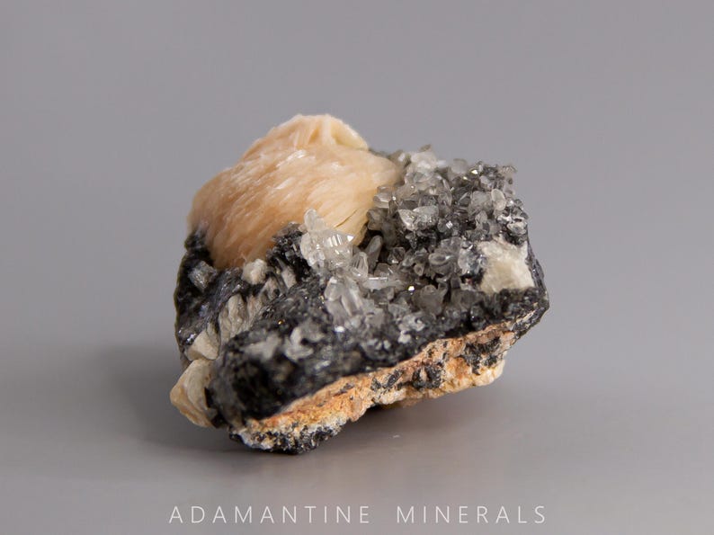 May include: A cluster of light pink barite crystals on a dark gray matrix. The barite crystals are translucent and have a pearly luster. The matrix is covered in small, clear quartz crystals.