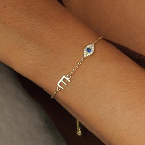 May include: Gold bracelet with an evil eye charm, featuring a blue centre and clear stone surround. The bracelet also has a small letter charm. A matching necklace with an evil eye pendant is also visible.