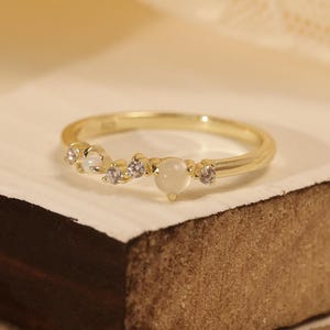 Könnte beinhalten: Ein goldener Ring mit filigranem Design. Der Ring hat eine Reihe kleiner, klarer Edelsteine und einen größeren, milchig-weißen Stein. Der Ring ist vor dem Hintergrund einer Holzoberfläche und weichem, cremefarbenem Stoff platziert.