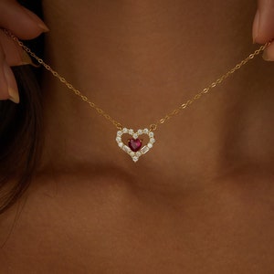 May include: A gold chain necklace with a heart-shaped pendant. The pendant is set with small clear stones and a larger red stone in the centre.