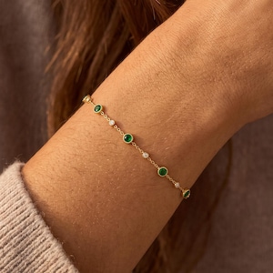 Könnte beinhalten: Ein zartes Goldkettenarmband mit abwechselnd runden grünen Edelsteinen und kleinen klaren weißen Steinen in Zargenfassung. Das Armband besteht aus einer dünnen metallischen Gliederkette, die sich um das Handgelenk legt und ein wiederkehrendes Muster zeigt.
