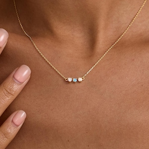 Peut inclure: Un collier à fine chaîne dorée orné de trois petites pierres précieuses rondes serties, disposées horizontalement au centre. Le bijou comprend deux pierres blanches transparentes sur les côtés et une pierre bleue irisée au milieu.