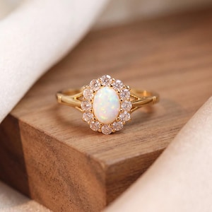 Pode incluir: Um anel de ouro com uma opala oval central rodeada por um halo de pequenas pedras transparentes de corte redondo. A banda é feita de ouro amarelo polido e tem um design de haste dividida que se conecta à cravação principal.
