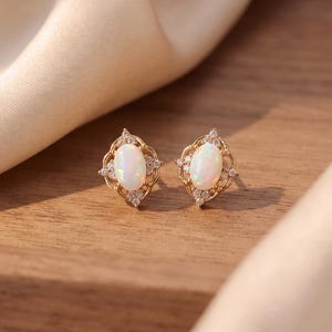 Könnte beinhalten: Ein Paar goldene Ohrstecker mit einem ovalen, schillernden Opal in der Mitte, umgeben von einem dekorativen filigranen Metallrahmen, der mit kleinen, klaren, runden Kristallen in einem symmetrischen Muster um den zentralen Opal verziert ist.