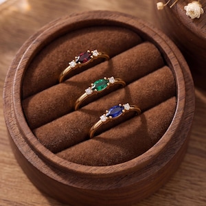 Puede incluir: Tres anillos de oro dispuestos en una caja de madera circular con forro de terciopelo marrón. Cada anillo presenta una piedra preciosa central de talla marquesa en rojo, verde o azul, flanqueada por dos pequeñas piedras claras.