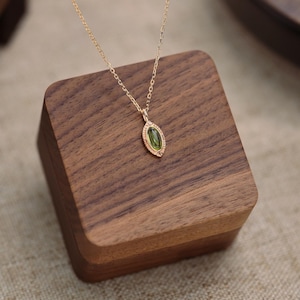 Könnte beinhalten: Eine zarte goldene Halskette mit einem kleinen, ovalförmigen, grünen Edelsteinanhänger. Der Anhänger ist in einen goldenen Rahmen gefasst und hängt an einer feinen Goldkette. Die Halskette wird auf einem kleinen, quadratischen Holzblock präsentiert.