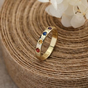 Op de afbeelding: Een gouden ring met stervormige zettingen, elk met een gekleurde edelsteen: rood, geel, blauw en paars. De ring is geplaatst tegen een achtergrond van opgerold touw en witte bloemen.