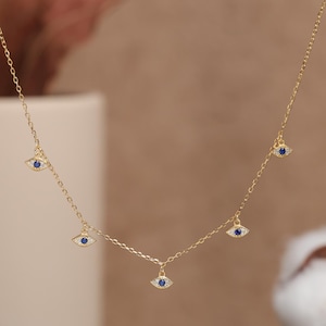May include: A delicate gold-colored necklace featuring multiple eye-shaped pendants. Each pendant has a blue center and is surrounded by small, clear stones. The necklace is displayed against a soft, neutral background.