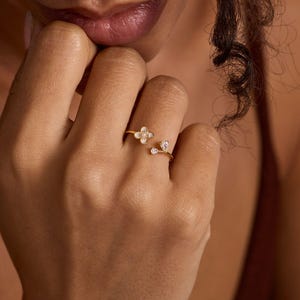 Könnte beinhalten: Ein goldfarbener offener Ring mit einem vierblättrigen Blumenmotiv mit weißer Mitte und einem separaten tropfenförmigen, klaren Steinakzent. Das Band ist dünn und metallisch und mit einem verstellbaren Spalt zwischen den Elementen gestaltet.