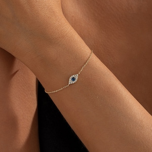 May include: A delicate gold chain bracelet featuring a central eye-shaped charm. The charm is set with a small, round, deep blue stone at the centre, surrounded by a halo of tiny, clear, sparkling white stones in a gold-toned metal setting.