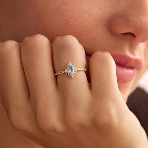 Könnte beinhalten: Ein Goldring mit einem birnenförmigen, hellblauen Edelstein in der Mitte, umgeben von einem zarten Filigranrahmen und kleinen, klaren Akzentsteinen. Die Ringschiene hat ein strukturiertes, gedrehtes Design, das zur Fassung hin schmaler wird.