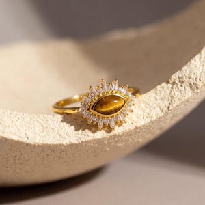 Pode incluir: Um anel de ouro com uma pedra central de olho de tigre oval, rodeada por um halo de pequenas pedras preciosas claras, cortadas em baguete e redondas. O anel tem uma banda fina e polida e uma cravação intrincada.