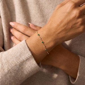 Puede incluir: Una delicada pulsera de cadena dorada con piedras preciosas redondas de color verde que se alternan con peque&ntilde;as piedras transparentes en engaste de bisel. La pulsera presenta una cadena de eslabones finos que rodea la mu&ntilde;eca, mostrando una disposici&oacute;n sencilla de piedras color esmeralda y destellos brillantes.