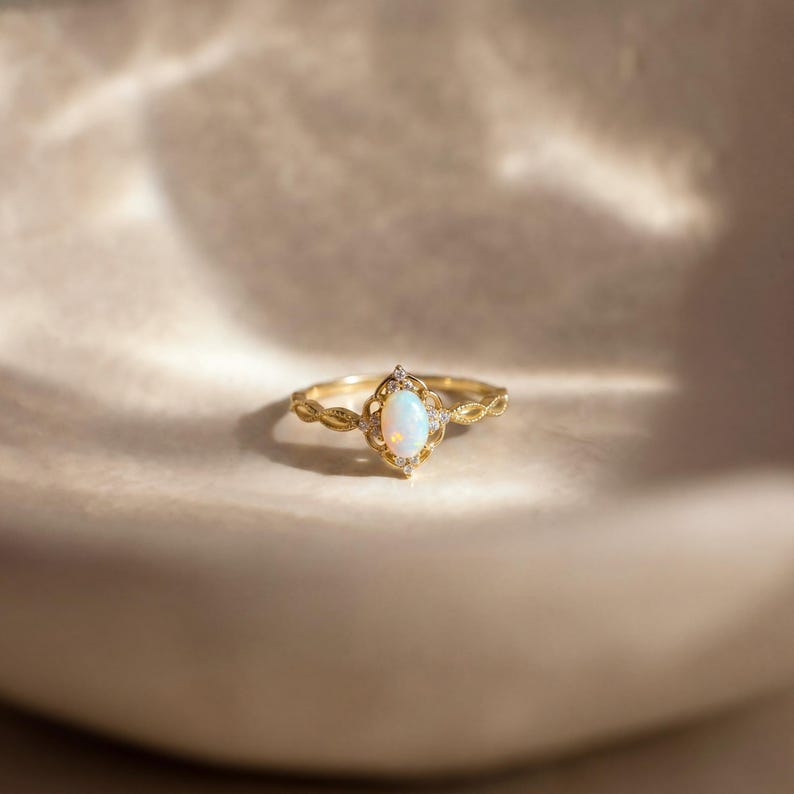 May include: A gold ring with an oval, iridescent opal center stone. The ring features a delicate, filigree design around the opal and along the band, accented with small, clear gemstones. The ring is set against a soft, neutral background.