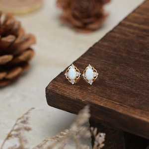 Op de afbeelding: Een paar goudkleurige oorstekers met ovale, witte opalen. Elke oorbel heeft een decoratief gouden filigraanontwerp en is versierd met kleine, heldere kristallen. De oorbellen worden gepresenteerd op een donker houten oppervlak.