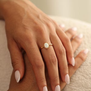 May include: A gold ring with an oval, white gemstone. The ring has a delicate, textured band and a scalloped setting around the gemstone. The ring is on a person's finger, with another hand in the background.