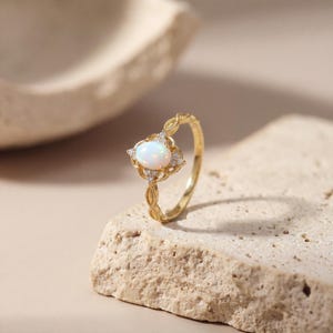 May include: A gold ring featuring an oval, white opal gemstone, surrounded by small, clear accent stones. The ring's band has a delicate, braided design. The ring is set on a textured, beige surface.