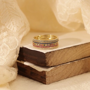 May include: A set of gold rings adorned with various coloured gemstones. The rings showcase rows of red, pink, blue, and green stones, interspersed with clear stones. They are presented on a small wooden surface with a white cloth.