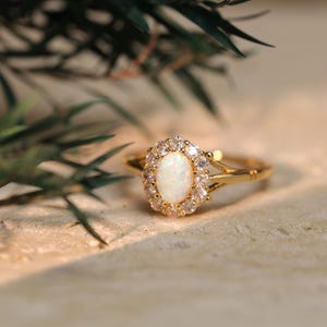 May include: A gold ring featuring an oval, white opal gemstone at its centre, encircled by a halo of smaller, clear stones. The ring's band has a delicate, split design. The ring is set against a neutral background with green foliage.