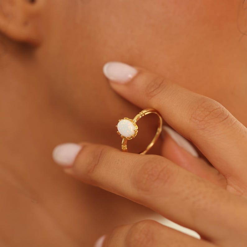 May include: A gold ring with an oval, white opal gemstone. The ring has a delicate, ornate design with a textured band and decorative accents around the stone. The ring is being worn on a finger.