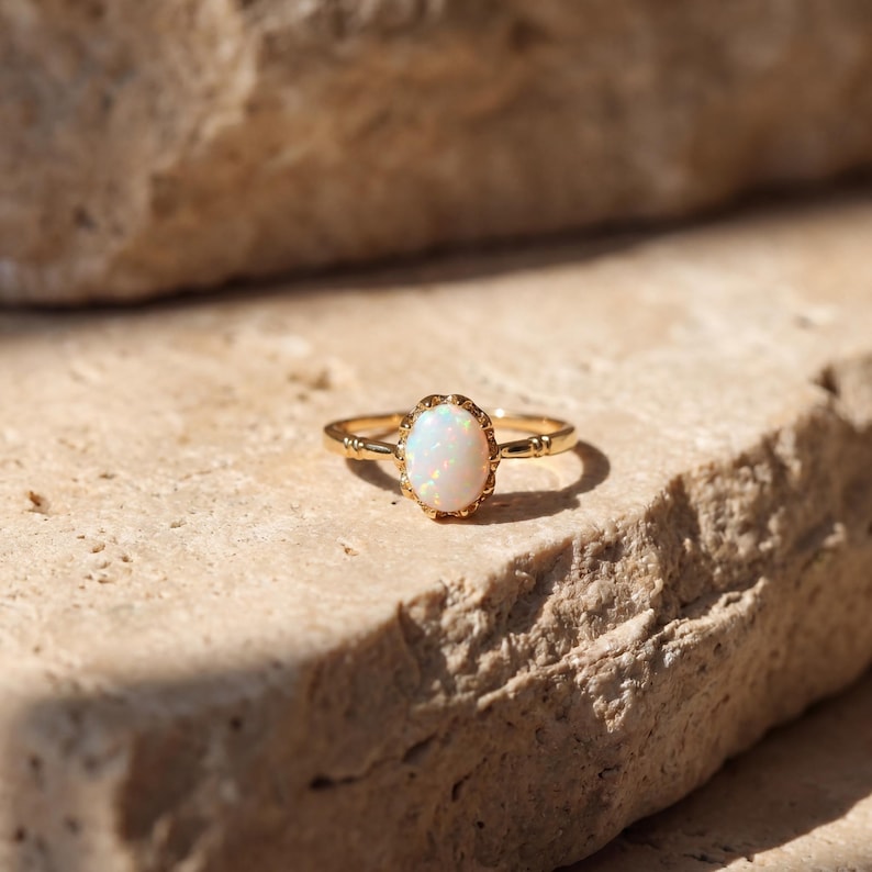 May include: A gold ring with an oval, white opal gemstone. The ring has a delicate, ornate design around the gemstone and along the band. The ring is set on a textured, light brown surface.